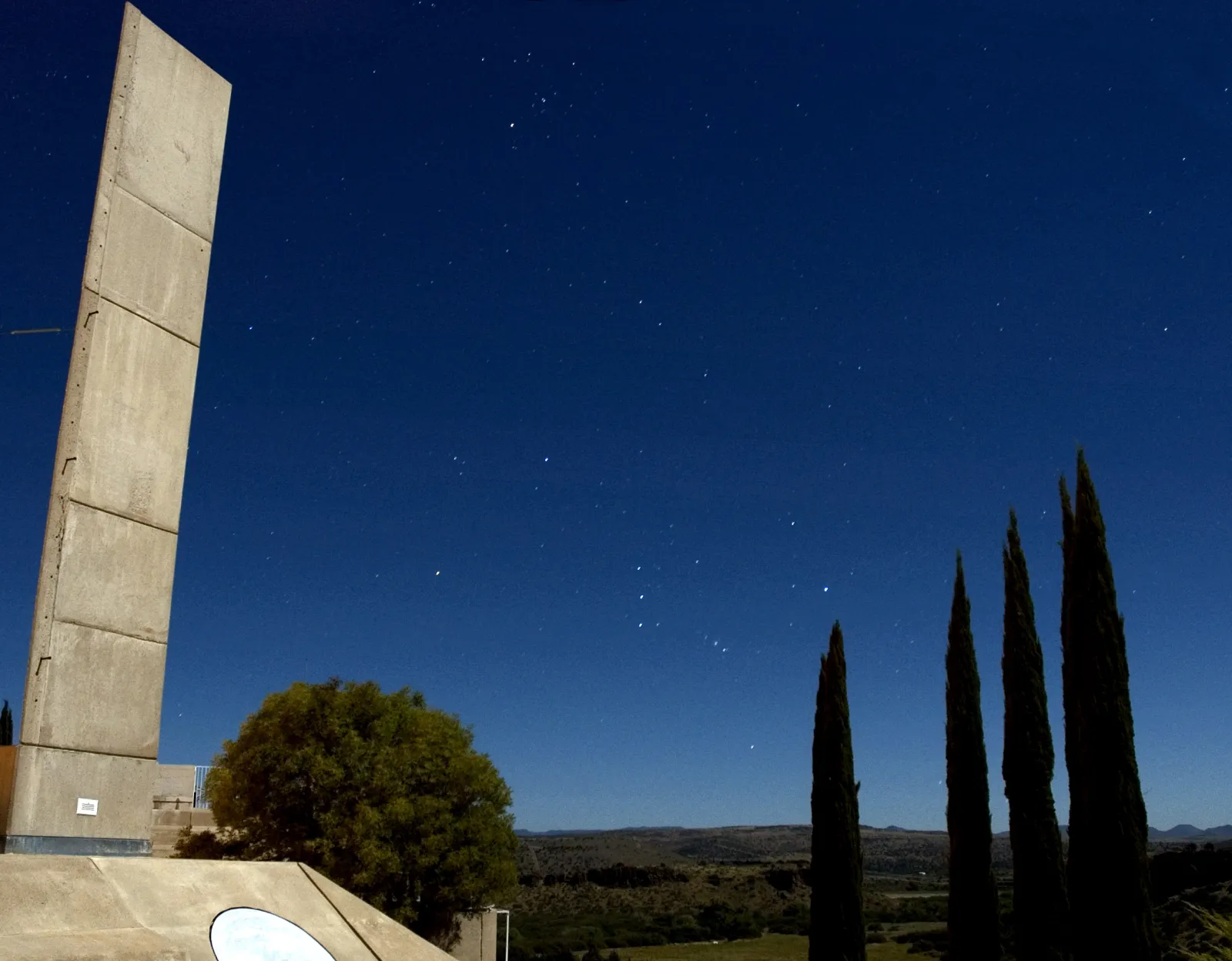 Orion rises between the cypress trees and concrete towers of Arcosanti at night